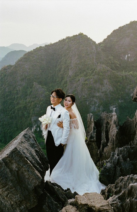 A couple in wedding attire stands on a rocky mountain peak with a bouquet of white flowers.