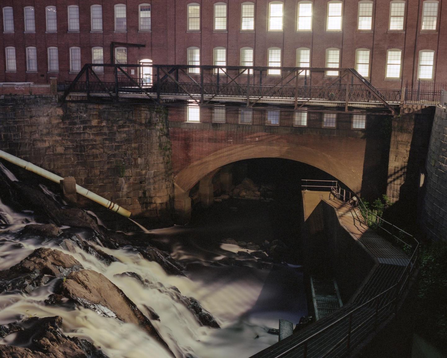 A waterfall flows under a bridge and a brick building with many illuminated windows at night.