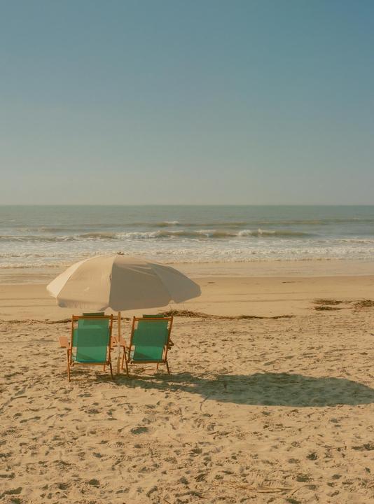 Two beach chairs sit under an umbrella on a sandy beach with the ocean in the background.