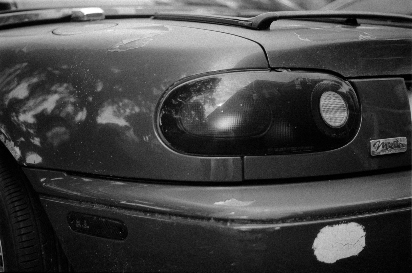 The rear of a dirty car with peeling paint is visible, showing the taillight and a "Miata" emblem.