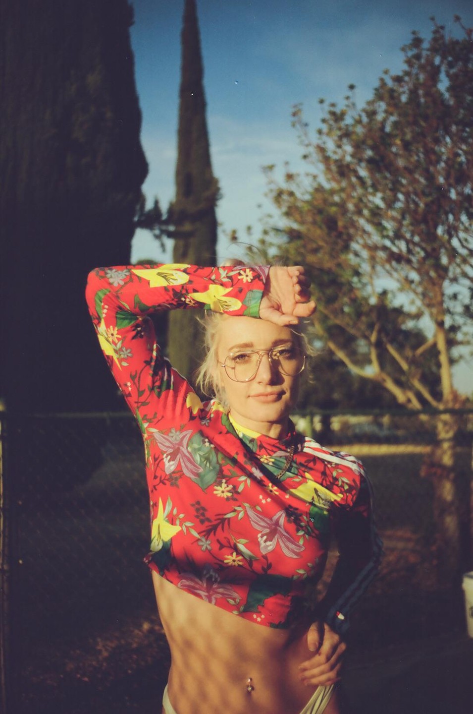 A woman in a red floral crop top and glasses poses with her arm raised to her forehead.