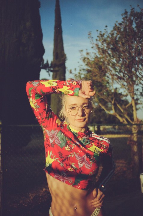 A woman in a red floral crop top and glasses poses with her arm raised to her forehead.