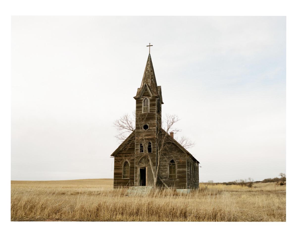 An old wooden church with a tall steeple stands in a field of dry grass under a pale sky.