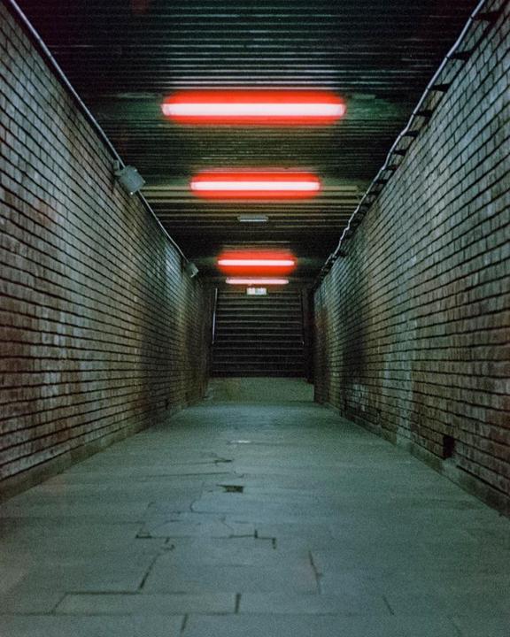 A long, narrow, brick-lined tunnel with red neon lights on the ceiling leads to a set of stairs.