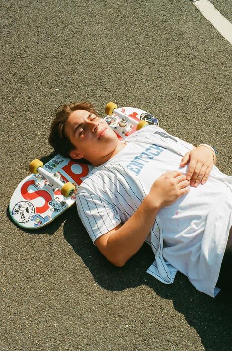 A young man with closed eyes is lying on a skateboard on an asphalt surface.
