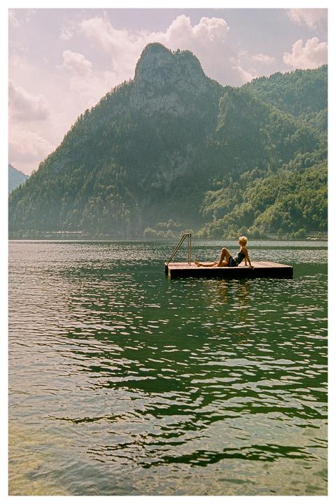 A woman in a swimsuit reclines on a floating dock in a lake surrounded by mountains.