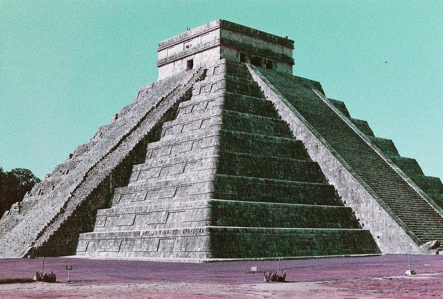 A large stone pyramid with multiple stepped levels and staircases rises under a clear sky.
