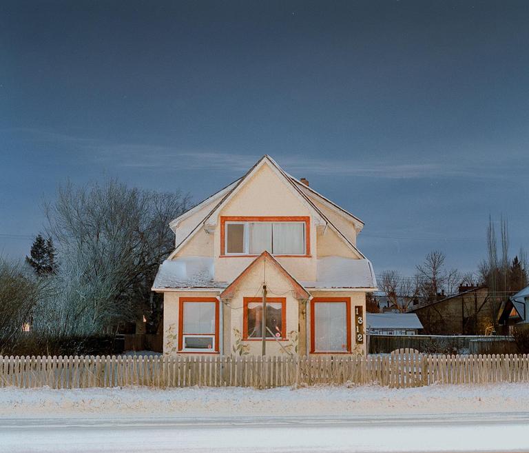 A cream-colored house with orange trim and a picket fence stands in a snowy landscape under a dark sky.