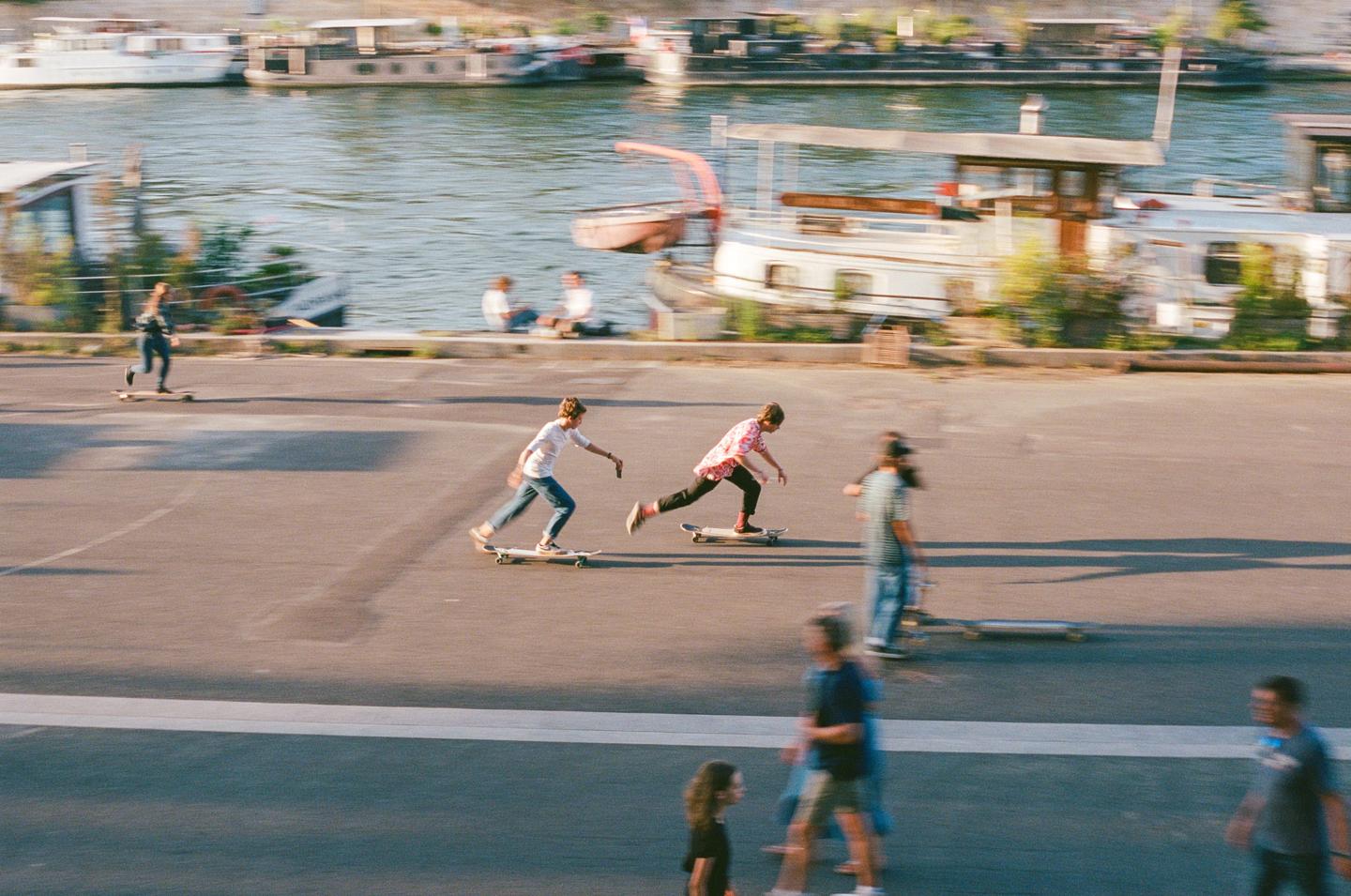 Two boys skateboard on a paved area next to a river with boats.