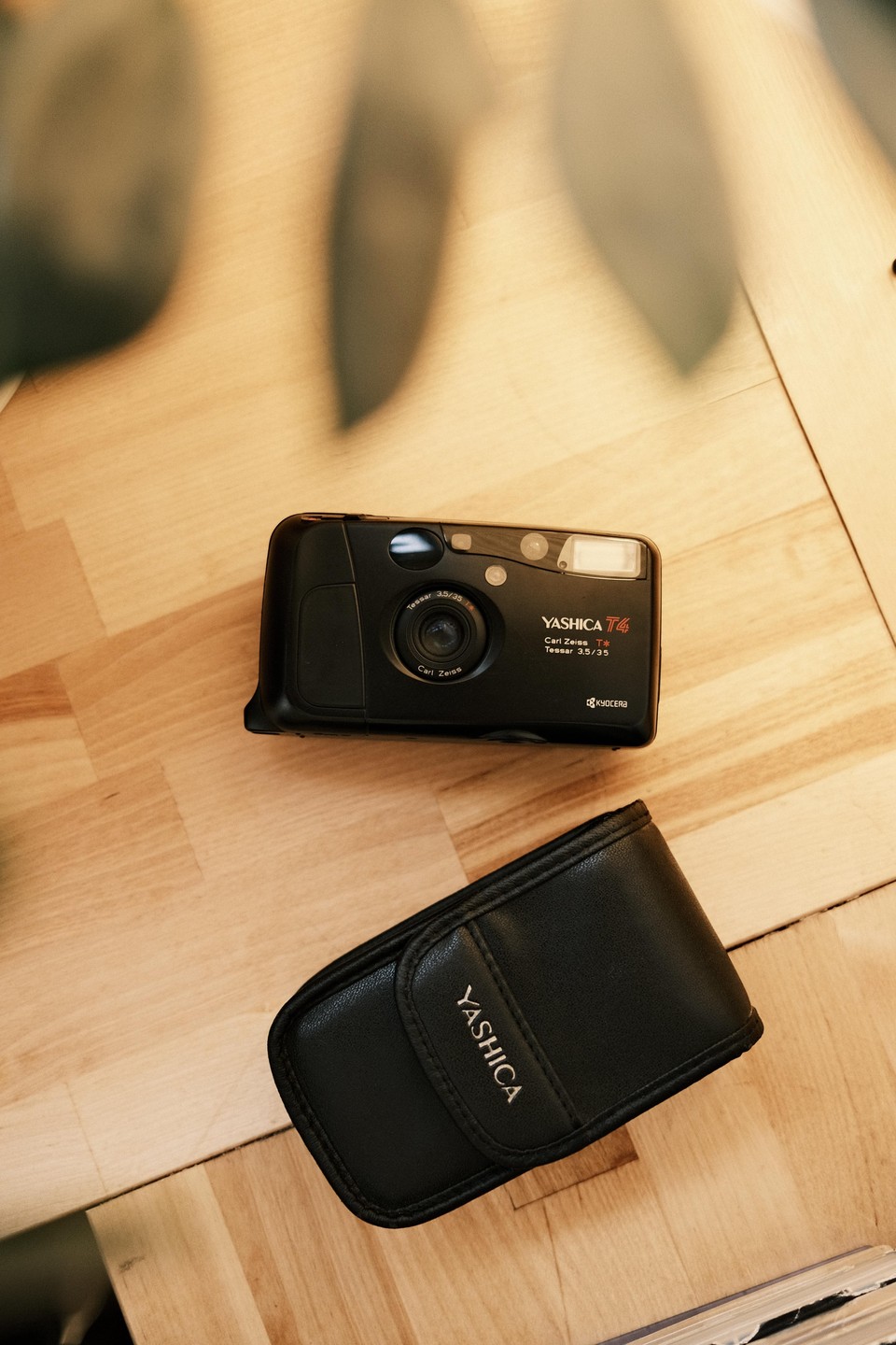 A black Yashica T4 camera and its black leather case are displayed on a wooden surface.