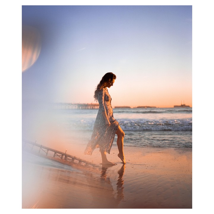 A woman in a long dress walks barefoot on a wet beach at sunset, with a pier and distant structures in the background.