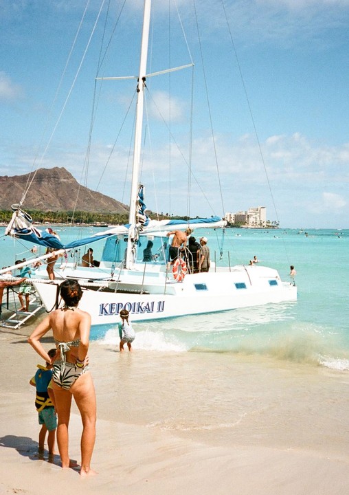 A woman and two children stand on a sandy beach next to a catamaran with people on board, with a mountain and buildings in the background.