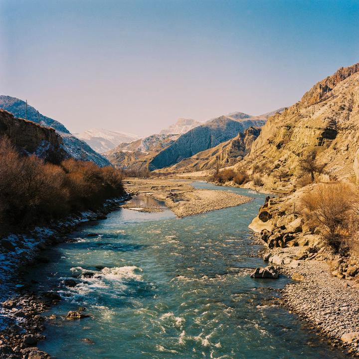 A river flows through a valley with mountains on either side, some of which are snow-capped.