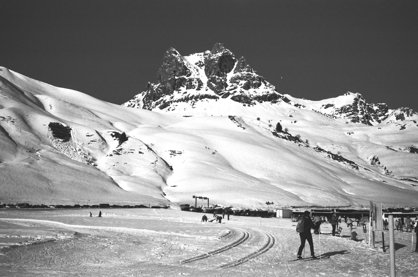 A person on skis stands in a snowy field with mountains in the background.