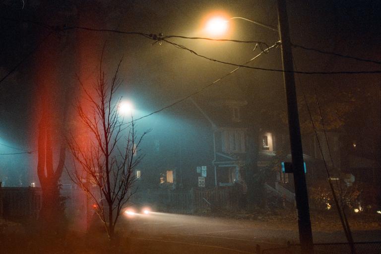 A street at night is illuminated by streetlights and car headlights, with a house visible through the fog.