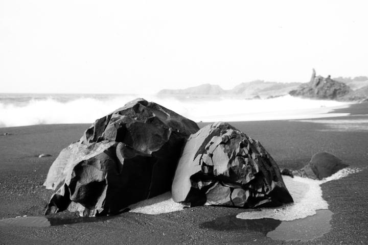 Two large rocks sit on a black sand beach with waves crashing in the background.