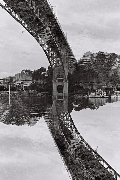 A double exposure shows a bridge over water with boats and houses on the shore, with the bridge reflected in the water.