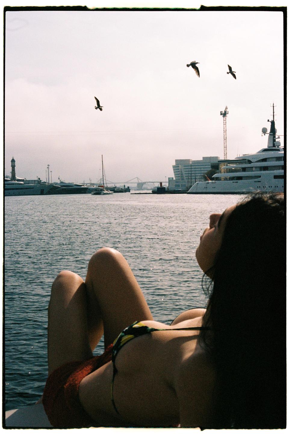 A woman in a bikini lies on a boat looking at the water with birds flying overhead and a city in the background.