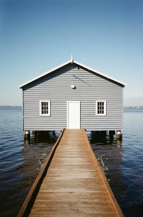 A wooden boardwalk leads to a blue boat house on stilts over calm water under a clear sky.