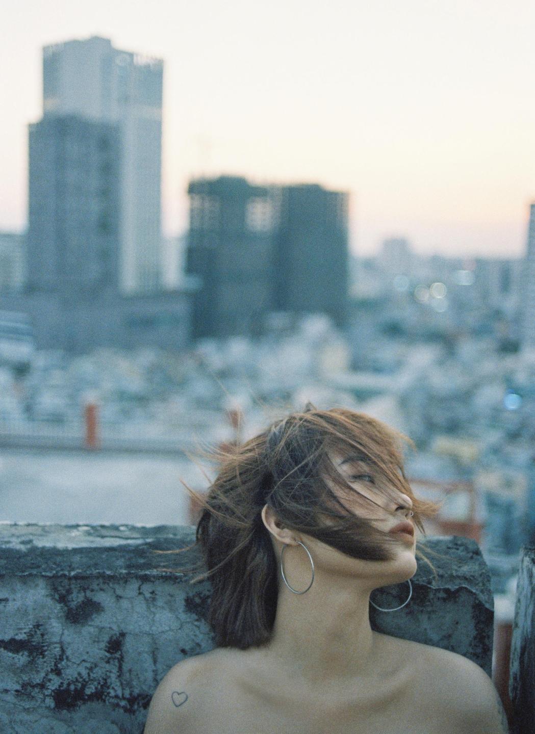 A woman with windblown hair and hoop earrings looks out over a city skyline at dusk.