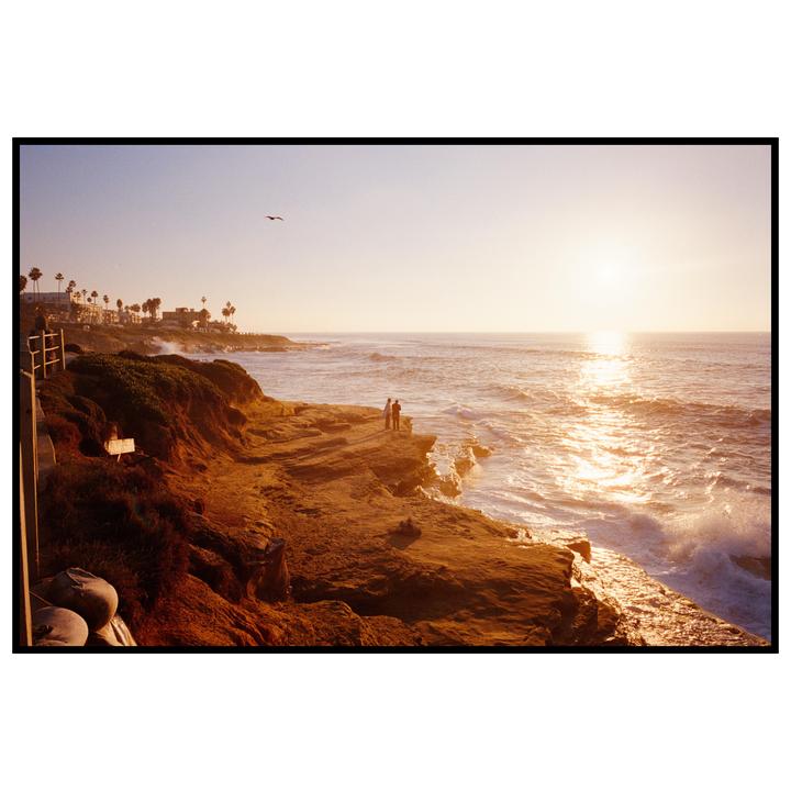 A couple stands on a rocky cliff overlooking the ocean at sunset.