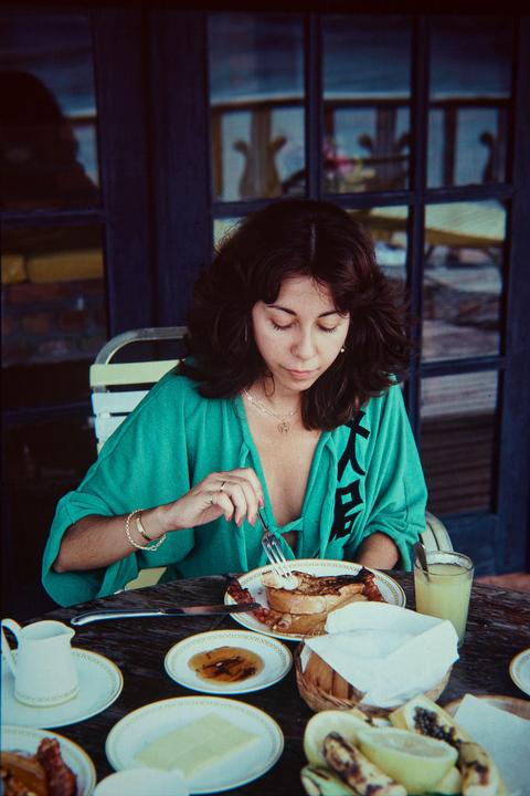 A woman in a green shirt eats breakfast at a table laden with food and drink.