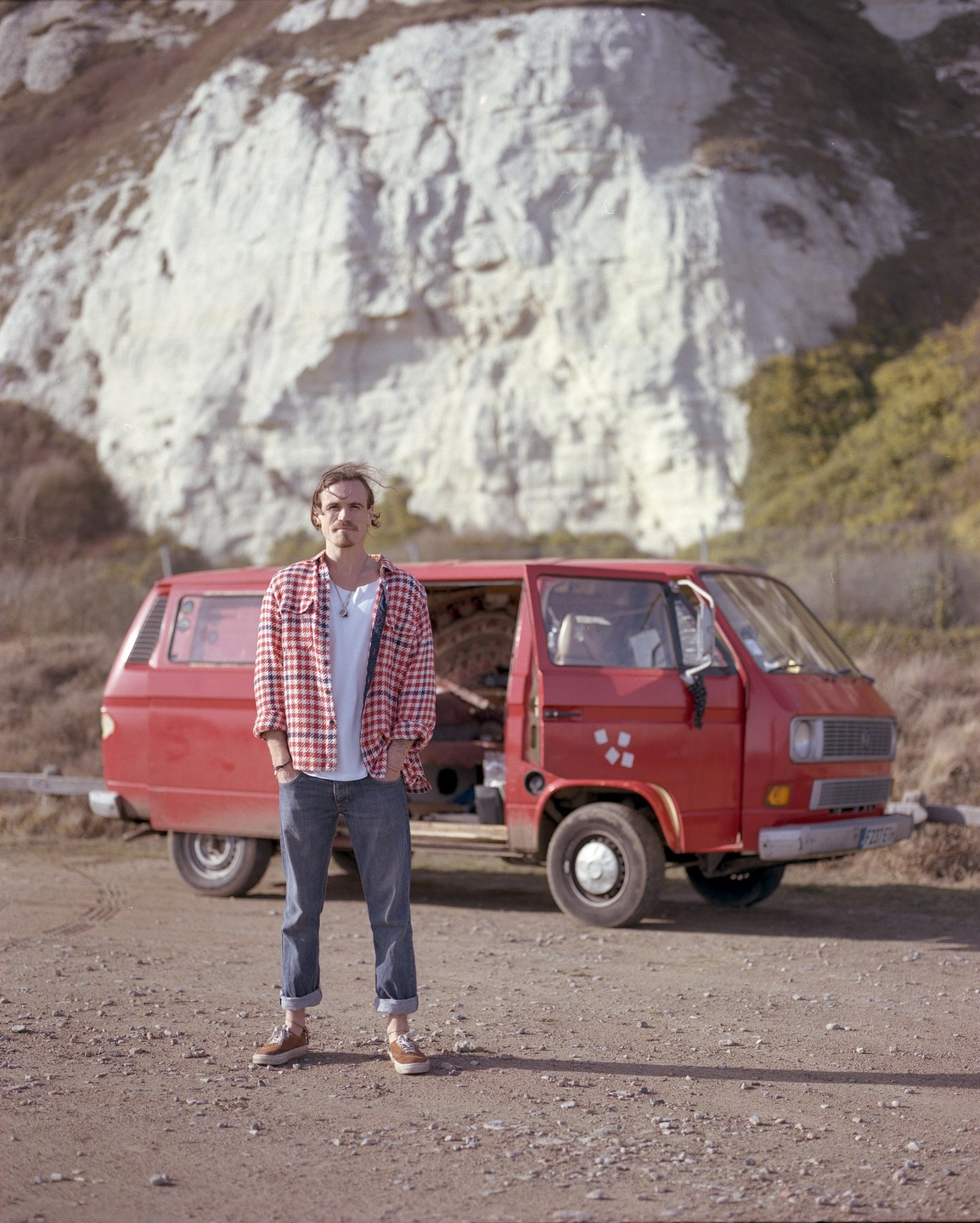 A man stands in front of a red van with a white cliff in the background.