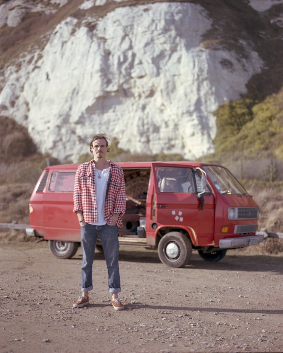 A man stands in front of a red van with a white cliff in the background.
