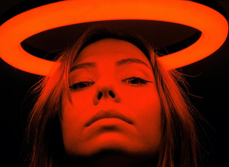 A woman's face is illuminated from below by a bright orange ring light that appears above her head.