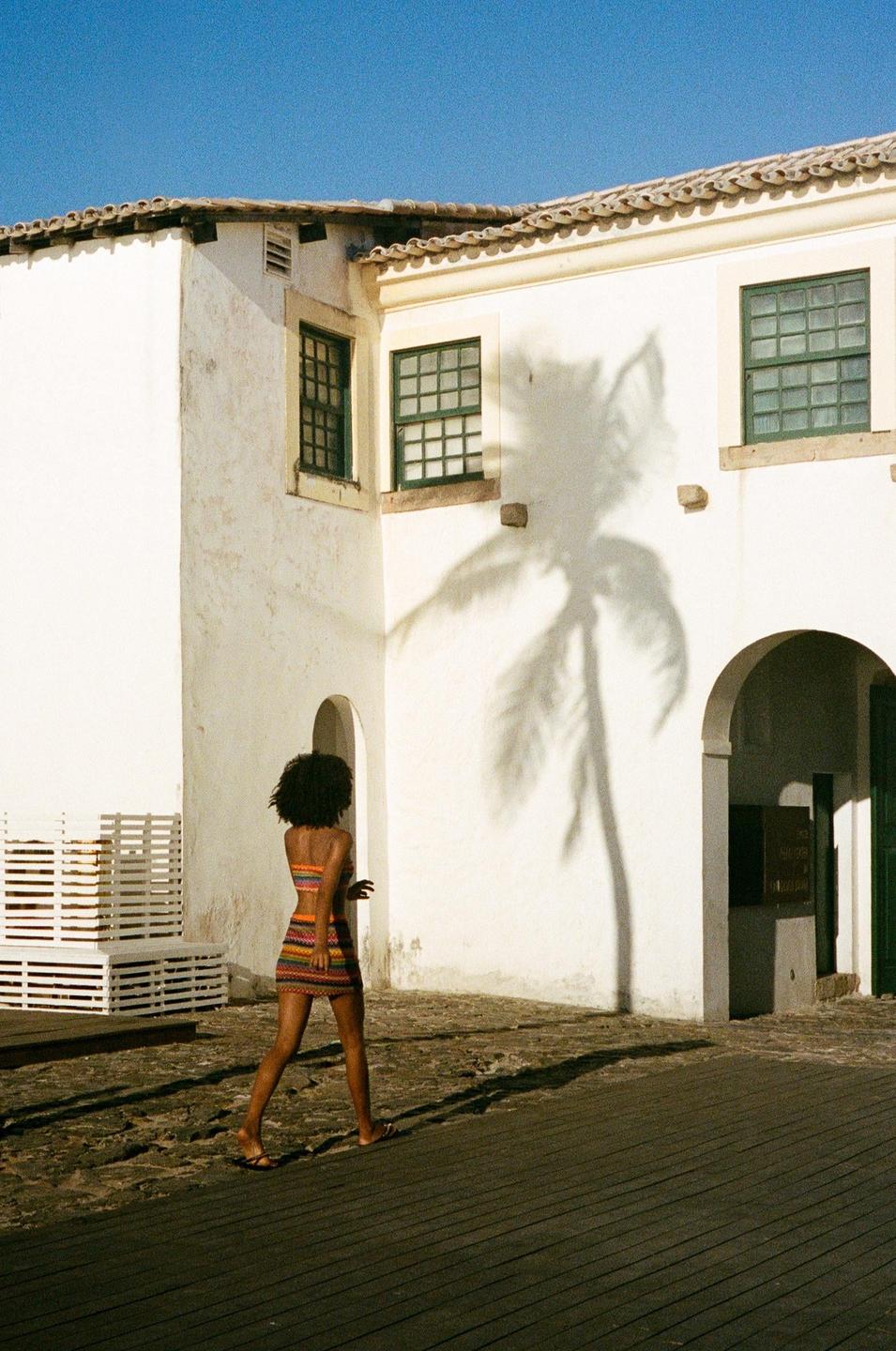 A woman in a colorful dress walks past a white building with a palm tree shadow cast on its wall.