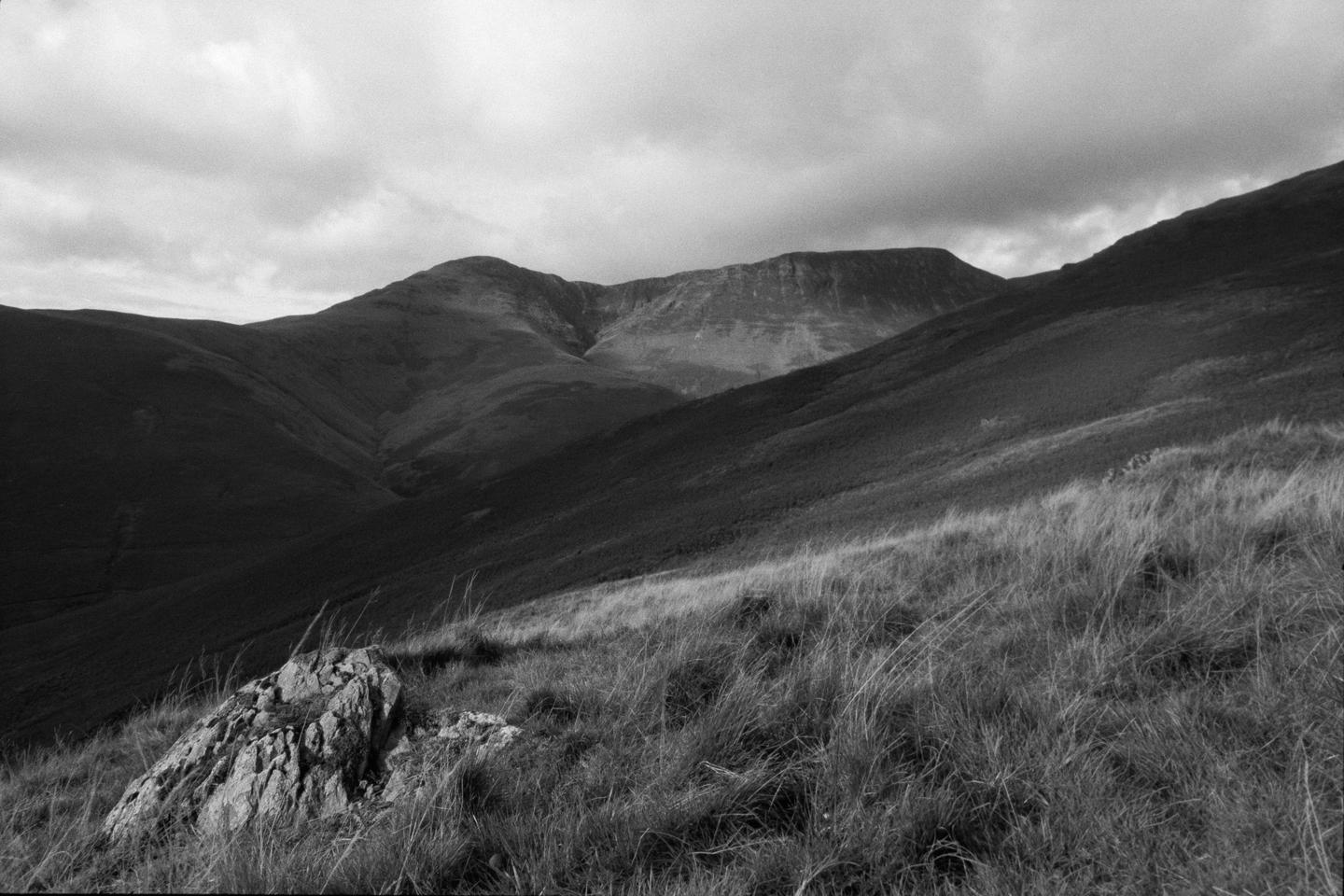 A black and white landscape shows a rocky, grassy hillside in the foreground and mountains under a cloudy sky in the background.