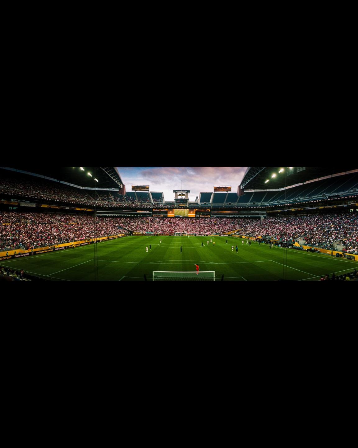 A panoramic view of a soccer match in a stadium filled with spectators under a cloudy sky.