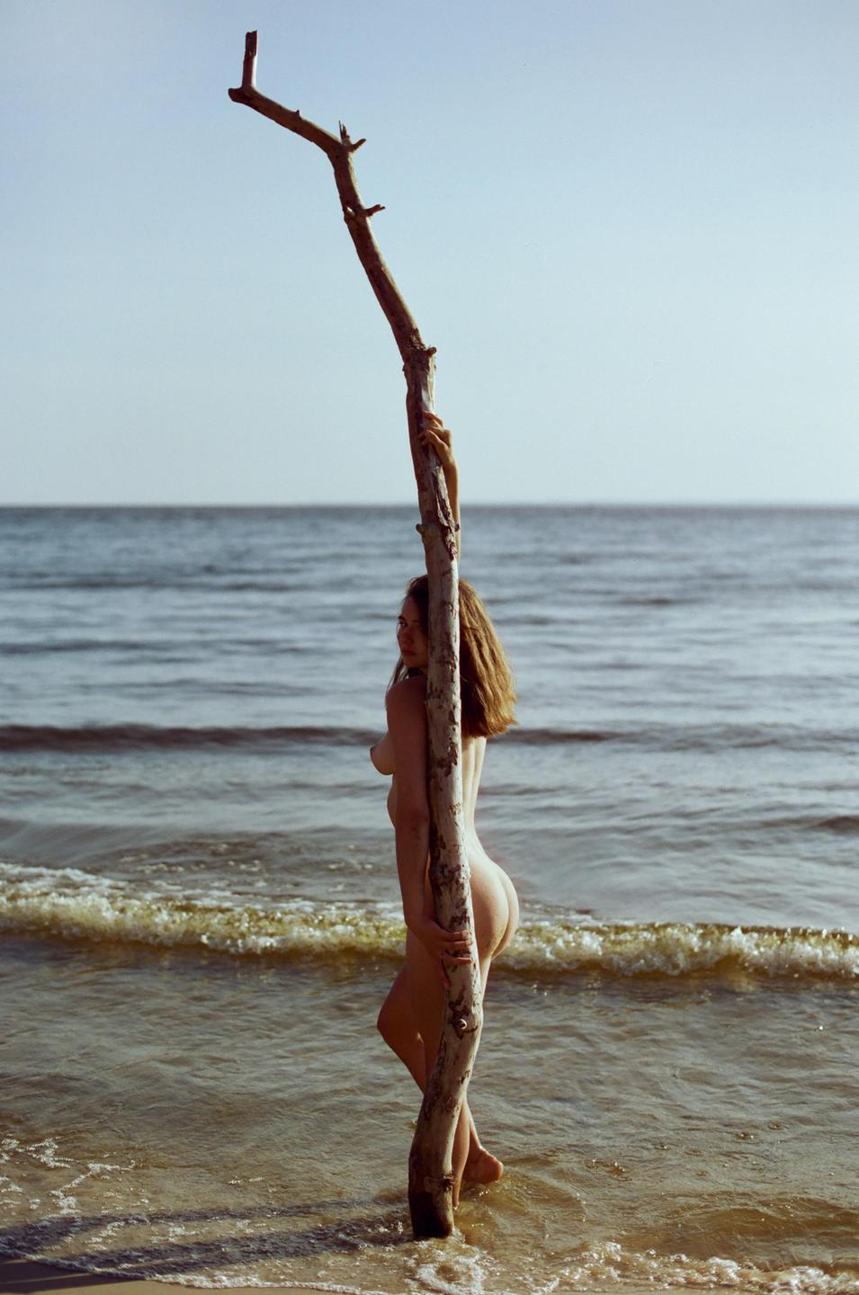 A nude woman stands in the ocean holding onto a large piece of driftwood.