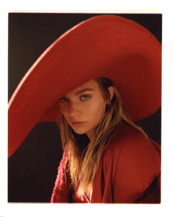 A woman wearing a large red hat and red clothing looks directly at the viewer.