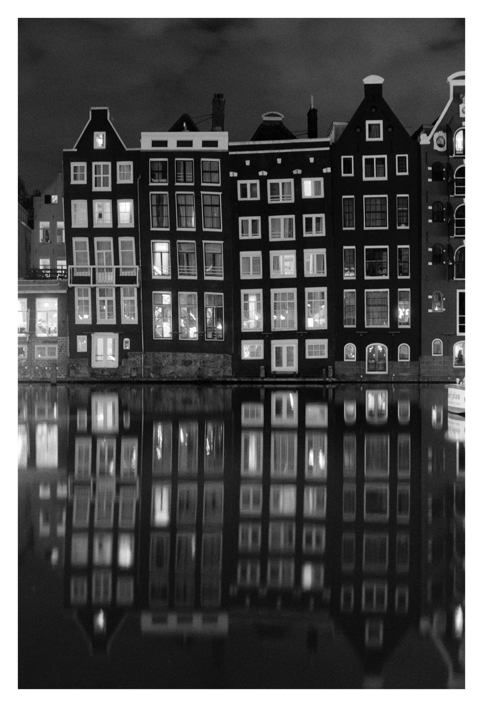 A row of tall, narrow buildings with many windows are reflected in the still water of a canal at night.