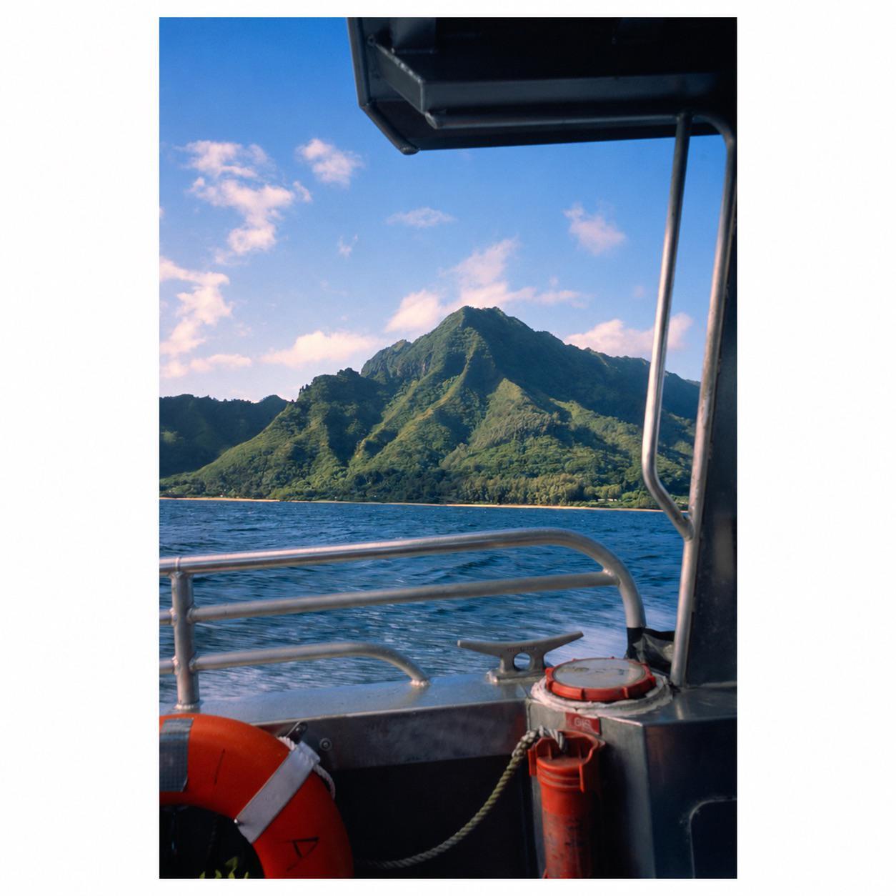 A boat on the water approaches a lush green mountain range with a small beach at its base under a blue sky with white clouds.