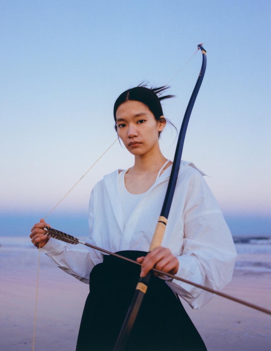 A woman holds a bow and arrow on a beach at dusk.