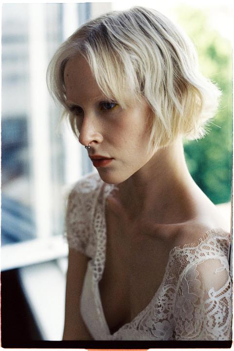 A woman with short blonde hair and a nose ring looks down, wearing a white lace top.