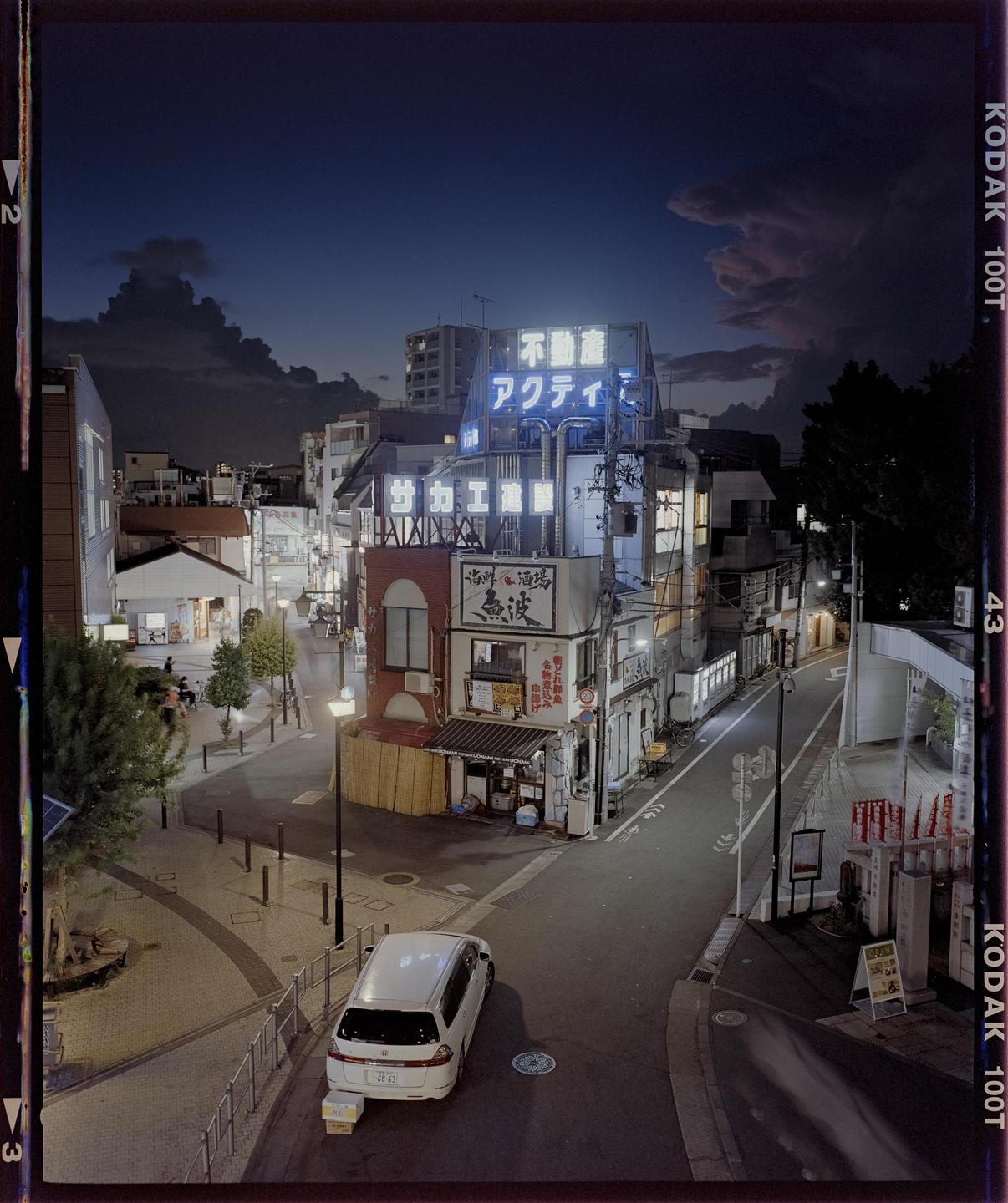 A street with buildings and a parked car is illuminated by neon signs and streetlights at dusk.