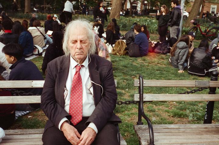 An elderly man with white hair and a red tie sits on a park bench with his eyes closed, wearing earphones, while other people relax on the grass in the background.