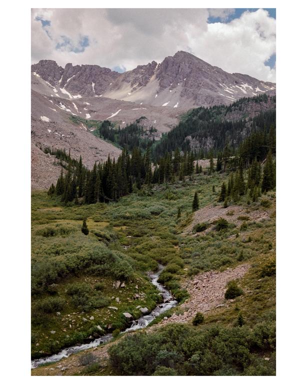 A stream winds through a valley with evergreen trees and mountains under a cloudy sky.