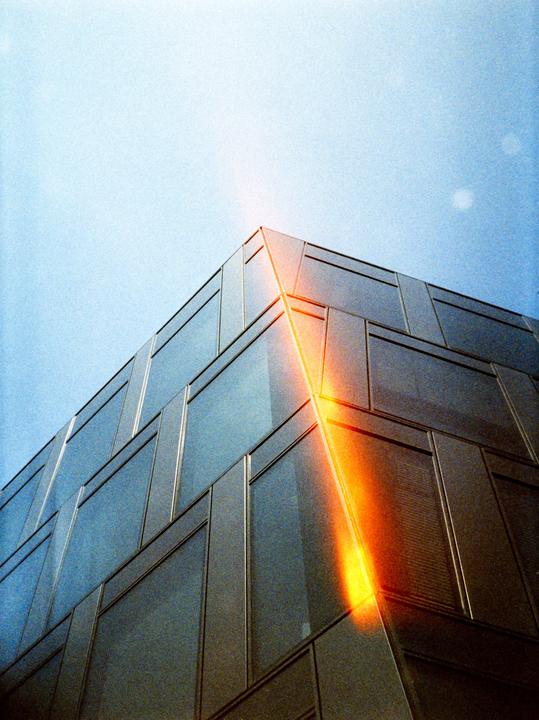A light leak illuminates the corner of a modern building with glass windows against a clear sky.