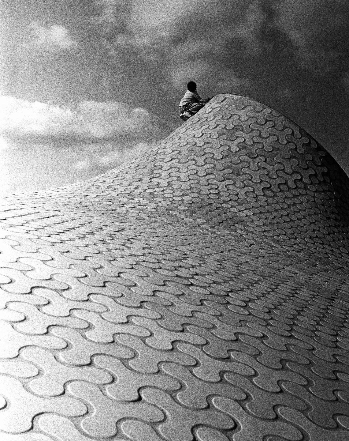 A person sits atop a patterned, undulating structure under a cloudy sky.