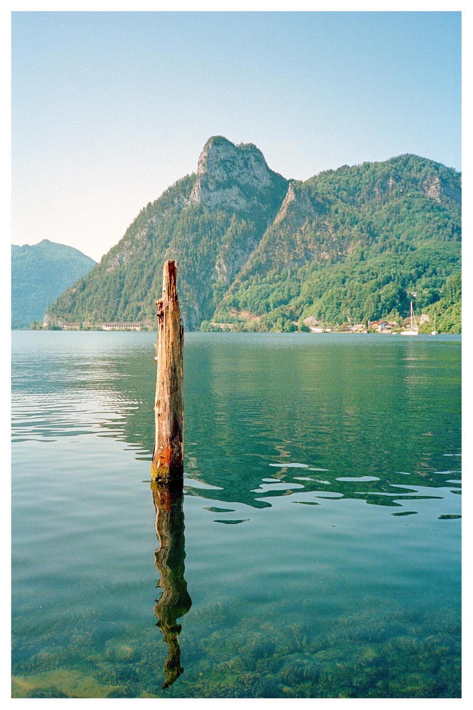 A wooden post stands in a lake with mountains and a small village in the background.