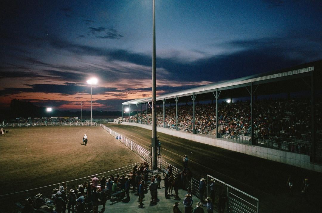 A cowboy on horseback is in the center of a rodeo arena with spectators in the stands under a dramatic sunset sky.