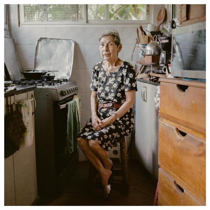 An elderly woman sits on a stool in a kitchen, looking directly at the viewer.