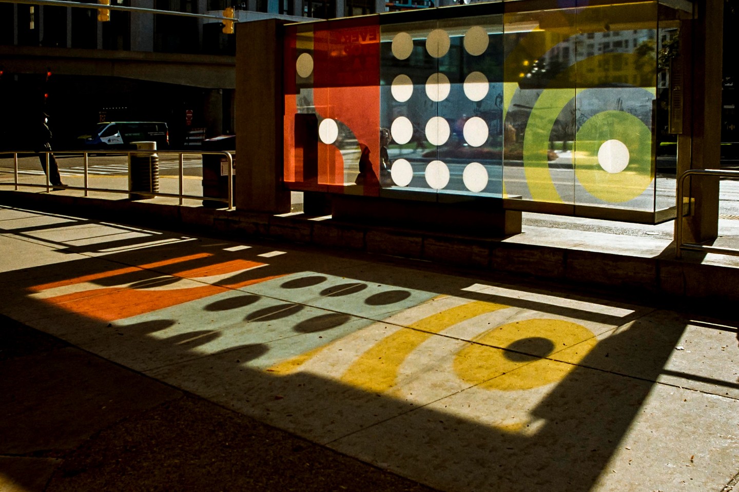 A bus stop with colorful panels casts shadows on the sidewalk, with a person standing nearby and a bus in the background.