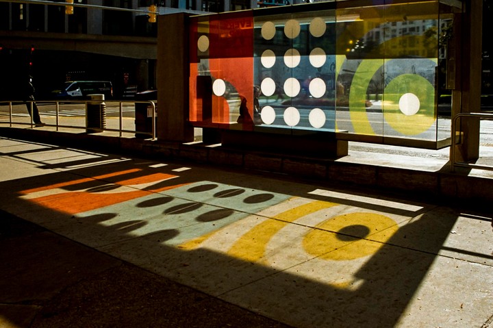 A bus stop with colorful panels casts shadows on the sidewalk, with a person standing nearby and a bus in the background.