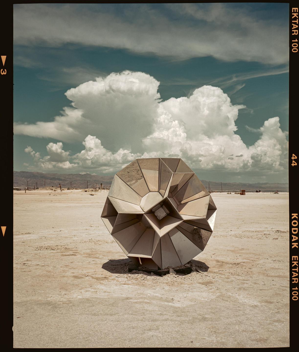 A geometric sculpture sits in a vast desert landscape under a cloudy sky.