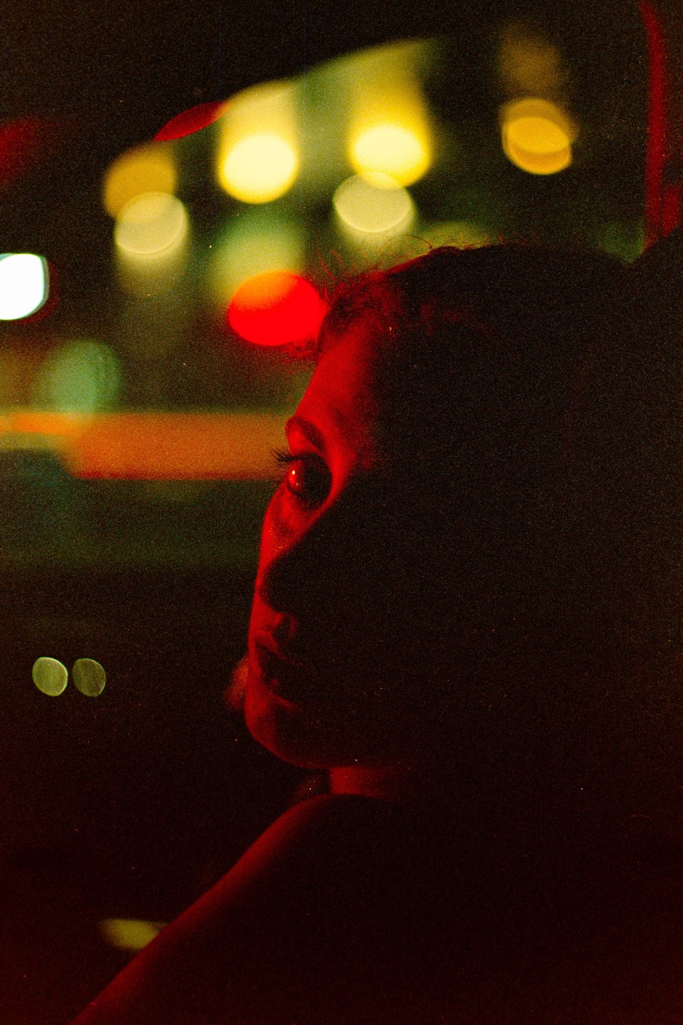 A woman's face is illuminated by red light in profile against a blurred background of colorful lights at night.